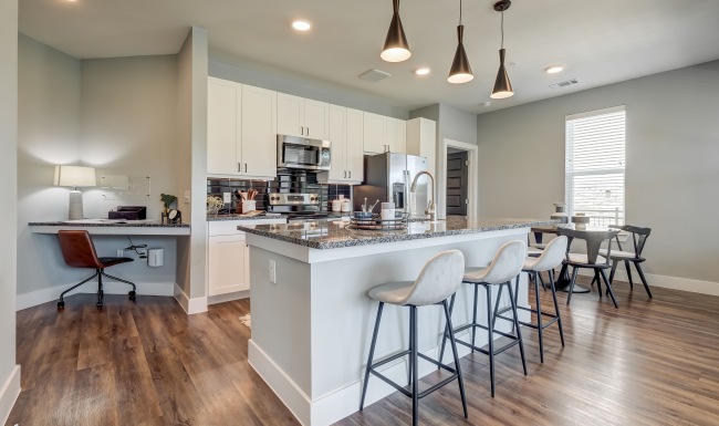 A modern kitchen with white cabinets, stainless steel appliances, a large island with granite countertops, barstools, pendant lights, and a small workspace with a desk and chair A modern kitchen with white cabinets, stainless steel appliances, a large island with granite countertops, barstools, pendant lights, and a small workspace with a desk and chair