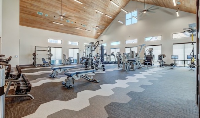 A spacious gym with high wooden ceilings, large windows, various exercise machines, free weights, and benches arranged on a geometric-patterned floor A spacious gym with high wooden ceilings, large windows, various exercise machines, free weights, and benches arranged on a geometric-patterned floor